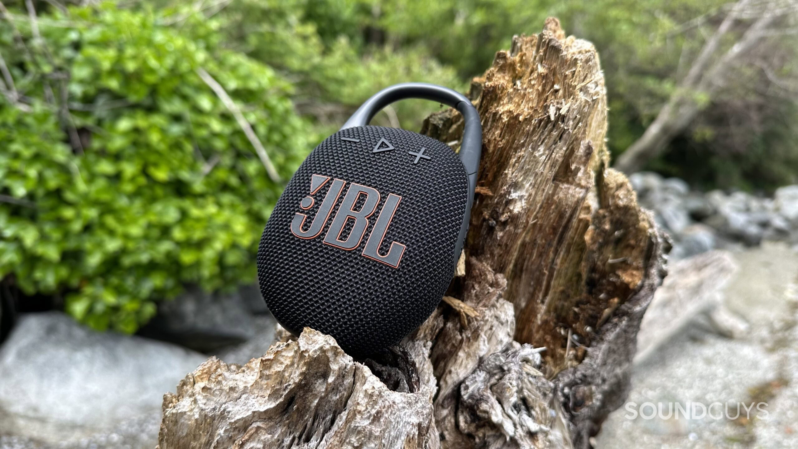 A JBL Clip 5 resting on a piece of beach driftwood.