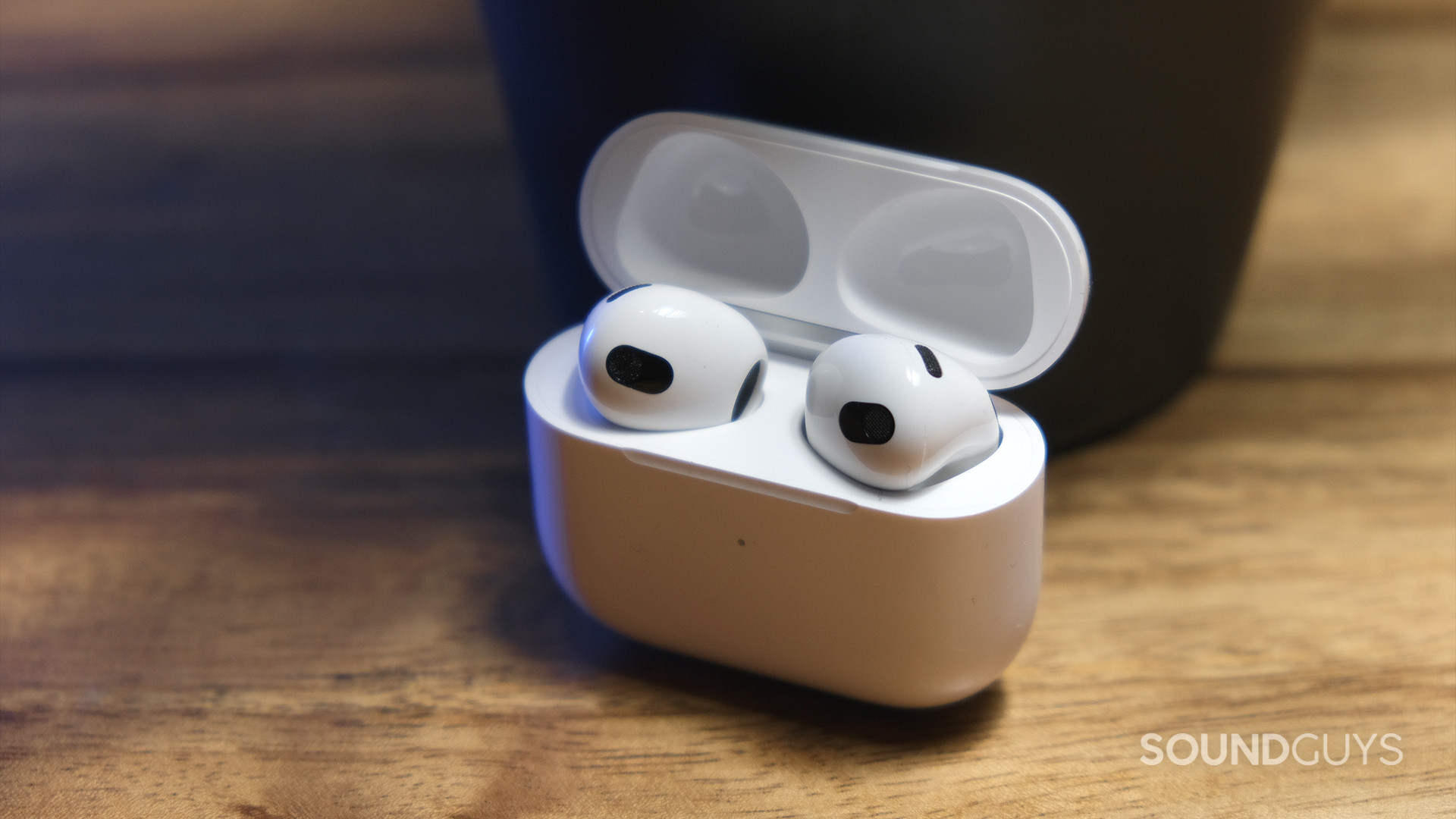 The Apple AirPods (3rd generation) open case holds the earbuds and sits on a wood surface.