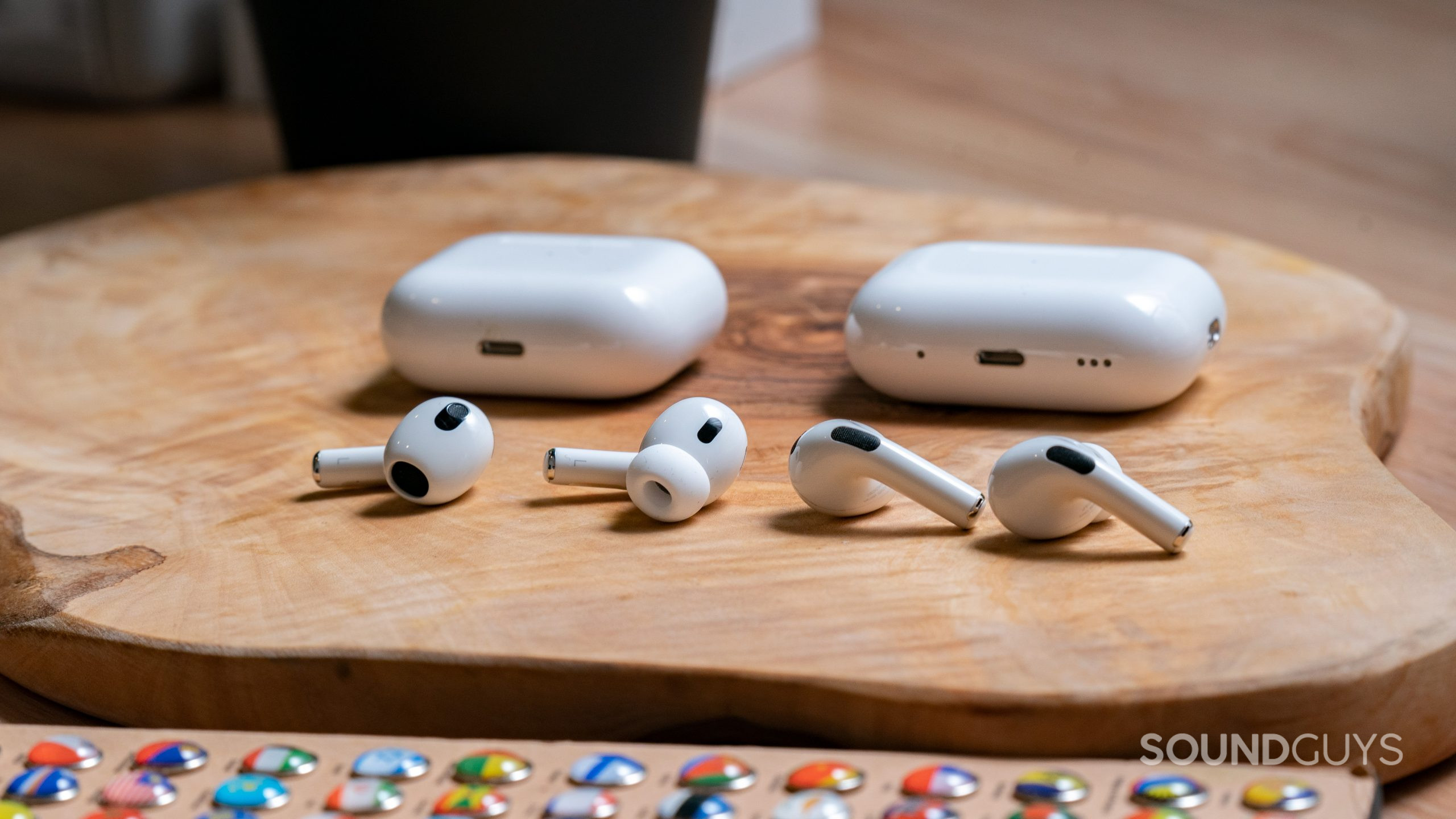 The Apple AirPods Pro Gen 2 and the Apple Airpods Gen 3 sitting on a wooden surface with their cases behind them.