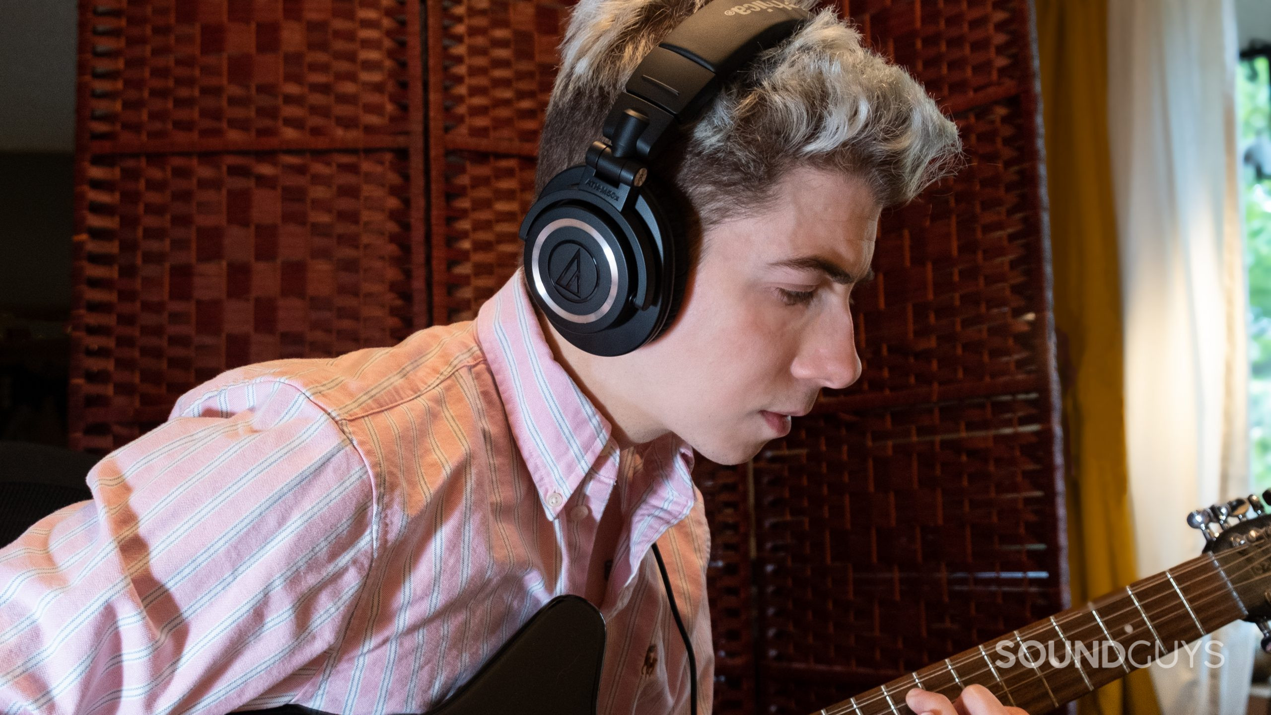 A man facing right plays guitar while wearing the Audio-Technica ATH-M50x.