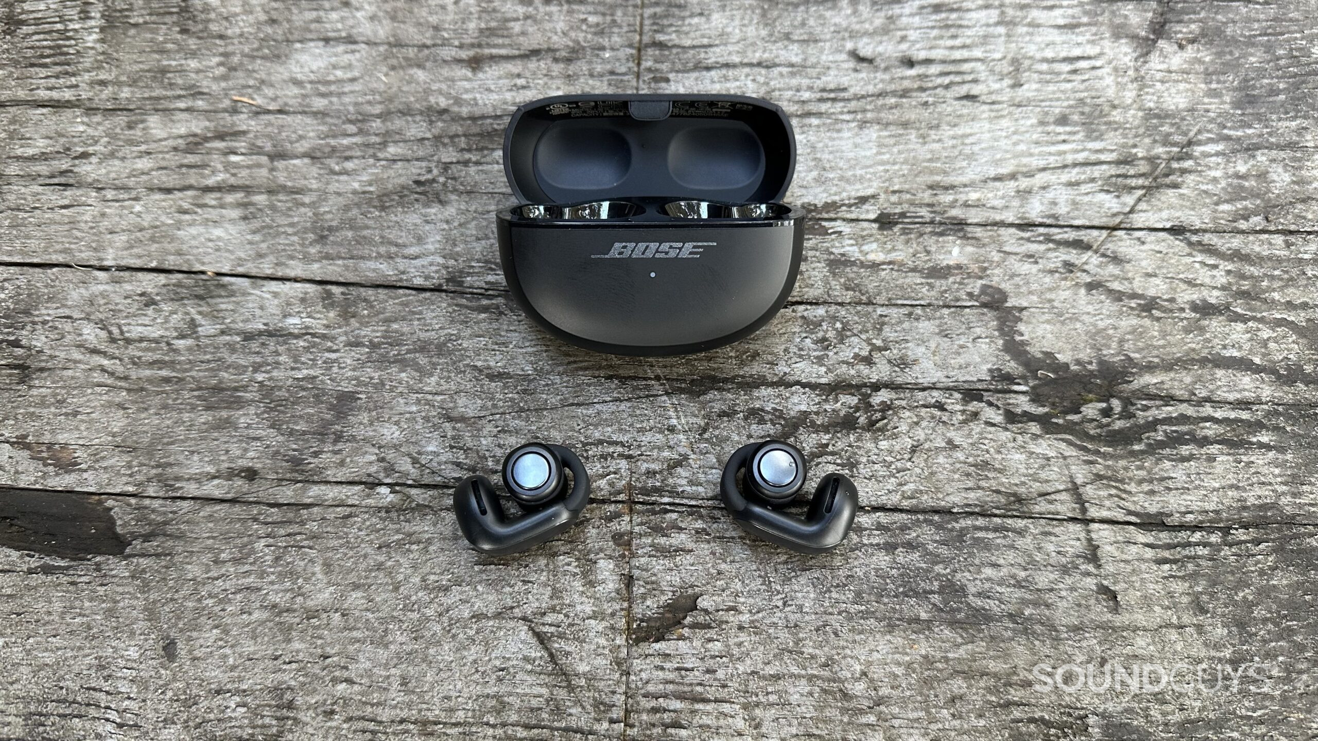 A pair of Bose Ultimate Open Earbuds and open charging case sitting on a grey bench