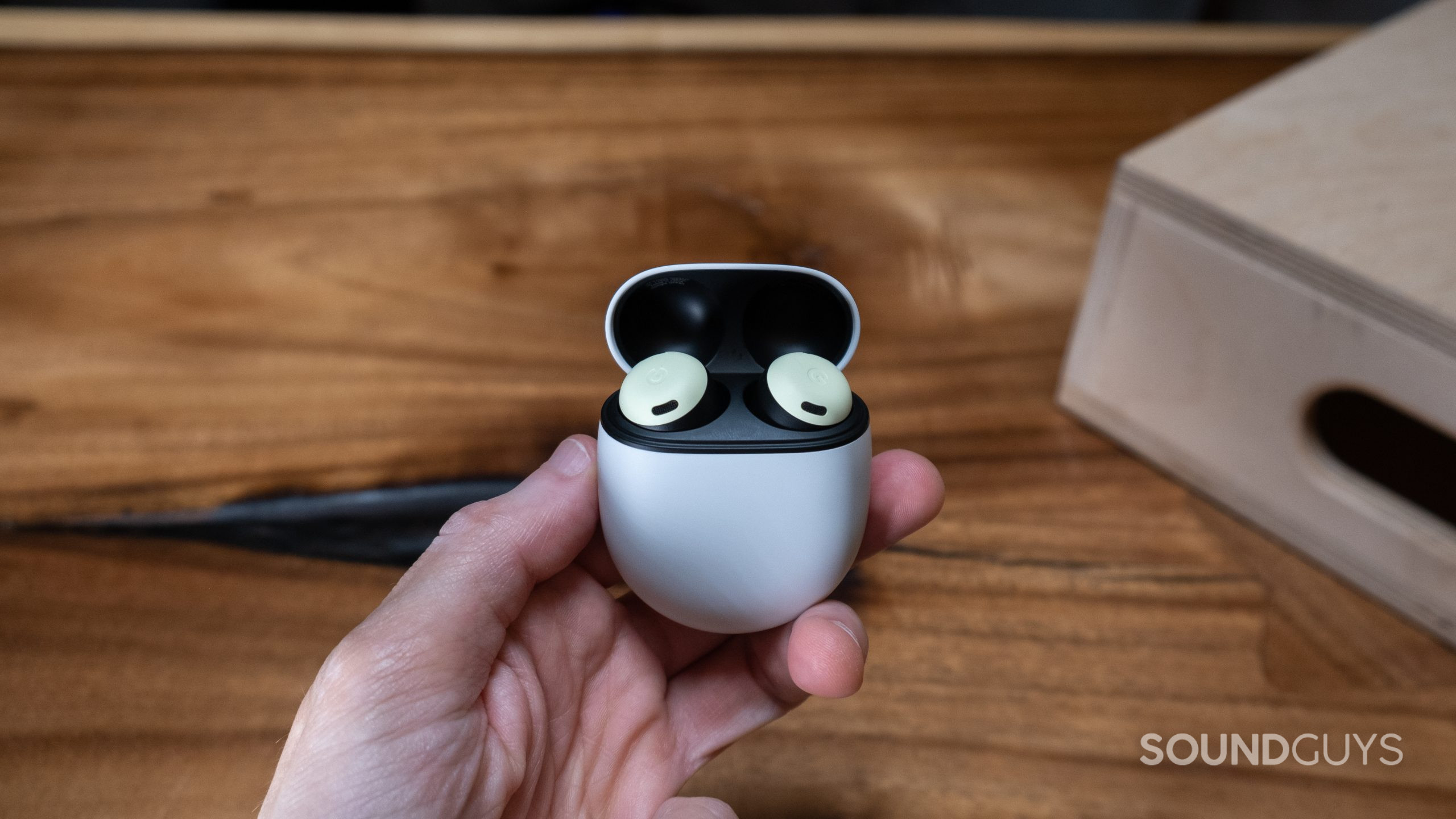 A hand holds the Google Pixel Buds Pro with the case open in front of a wood surface.