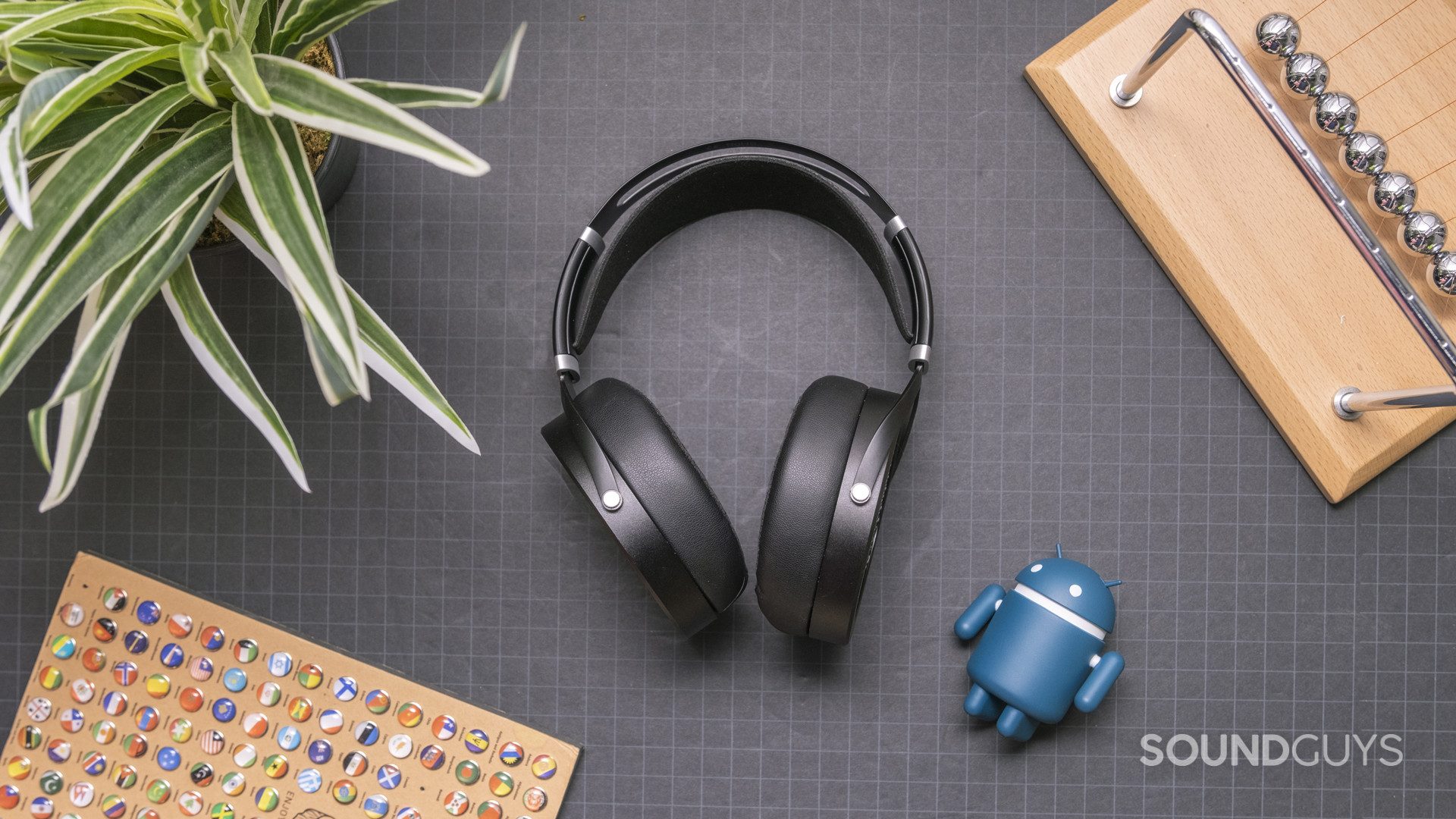 The HiFiMan Sundara rests on a gridded black surface with an artificial plant, Android toy, and wood accessories in the background.