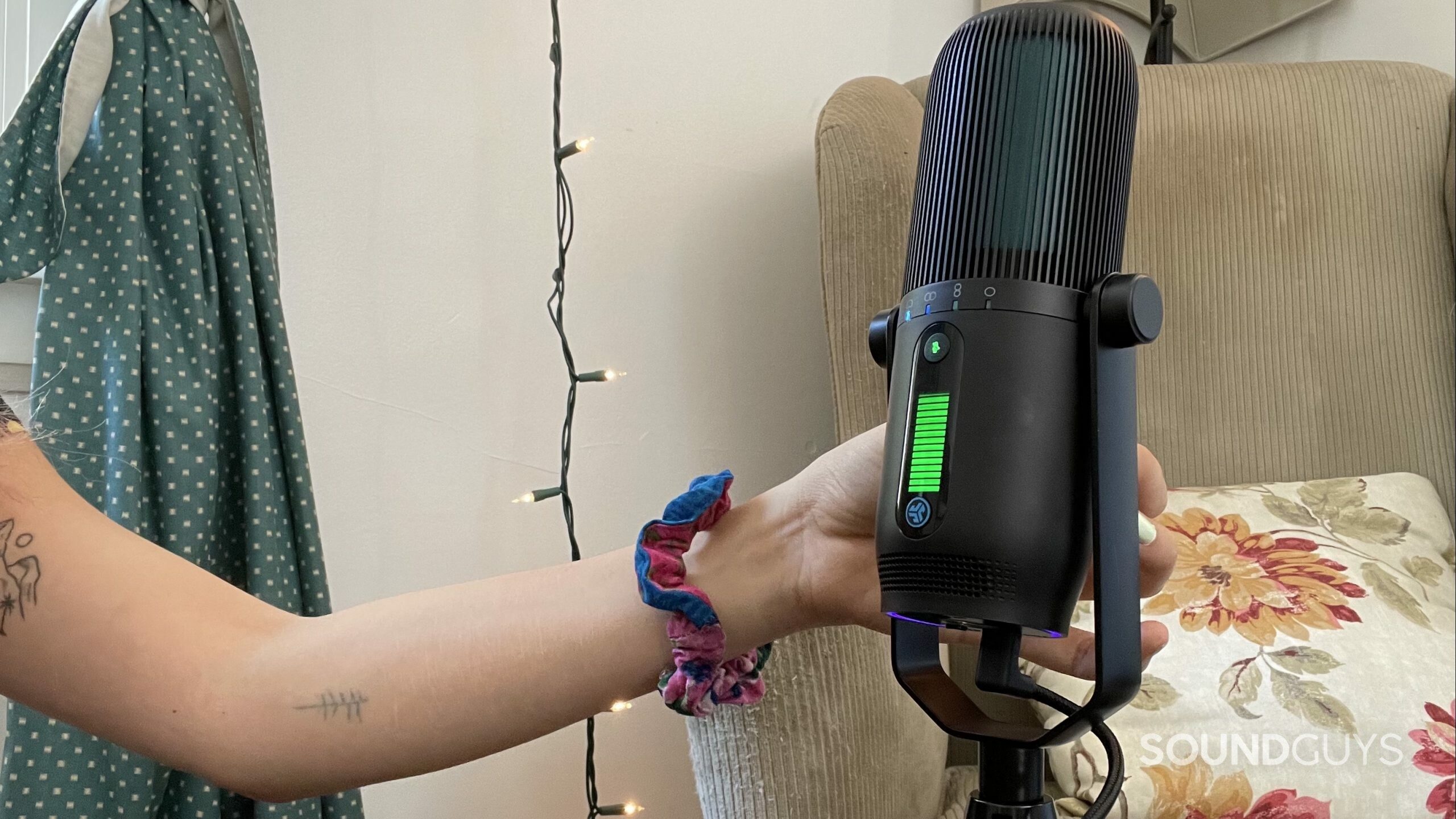 A womans hand using the JLab Talk PRO USB microphone with an armchair in the background.