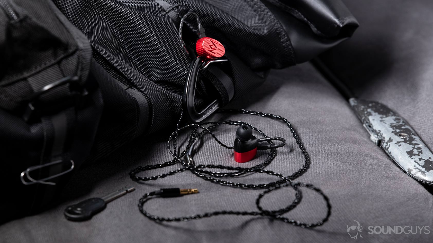 A level photo of the Massdrop x Noble Kaiser 10 Universal earbuds hanging off of a black back pack with a box cutter to the right side of the image.