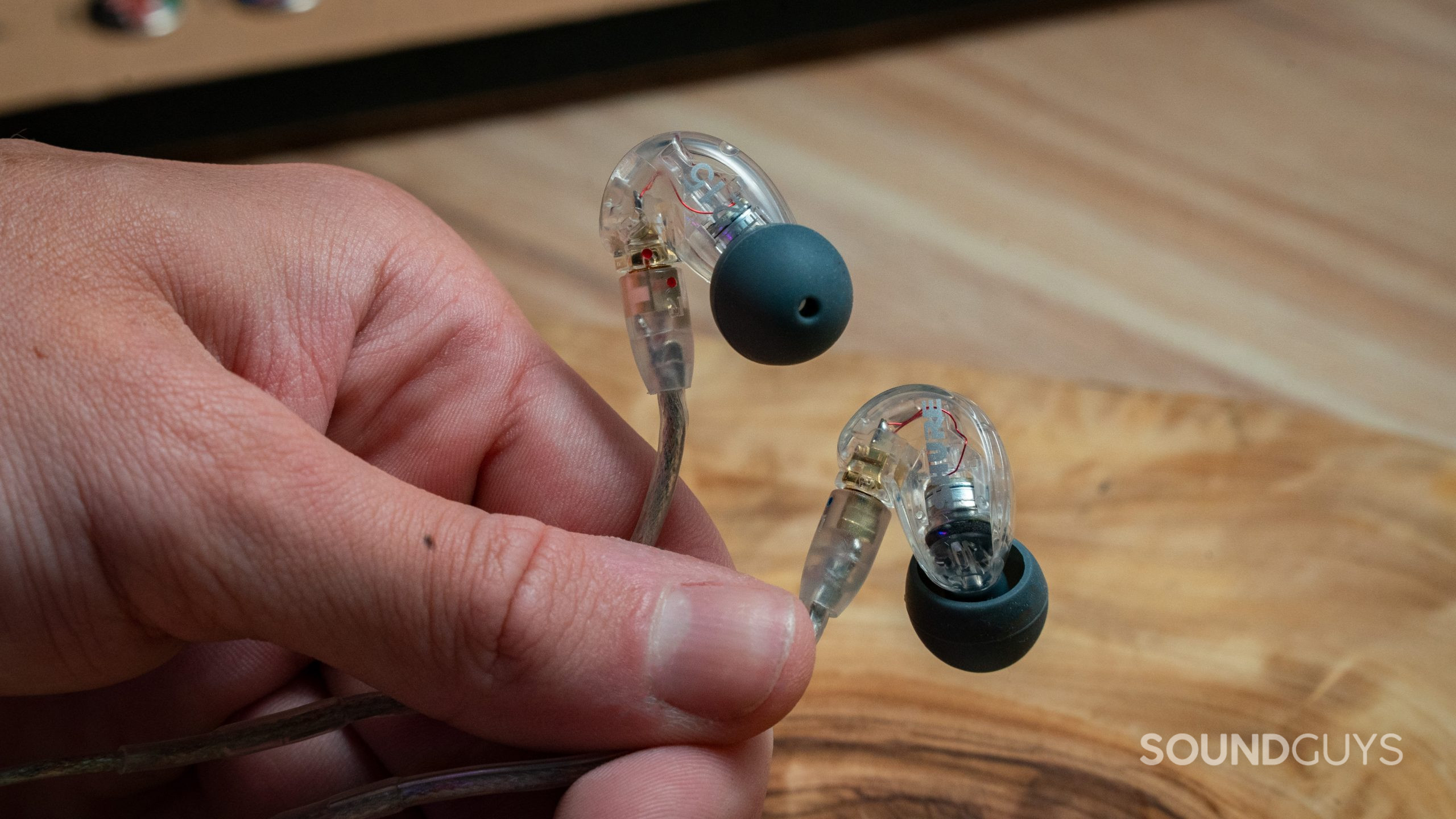 A hand holds the Shure SE215 in front of a wood table.