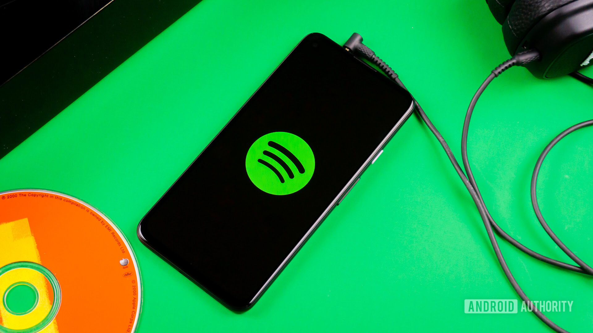 A phone sits on a green background, plugged into a set of wired earbuds. On the screen is the Spotify logo.